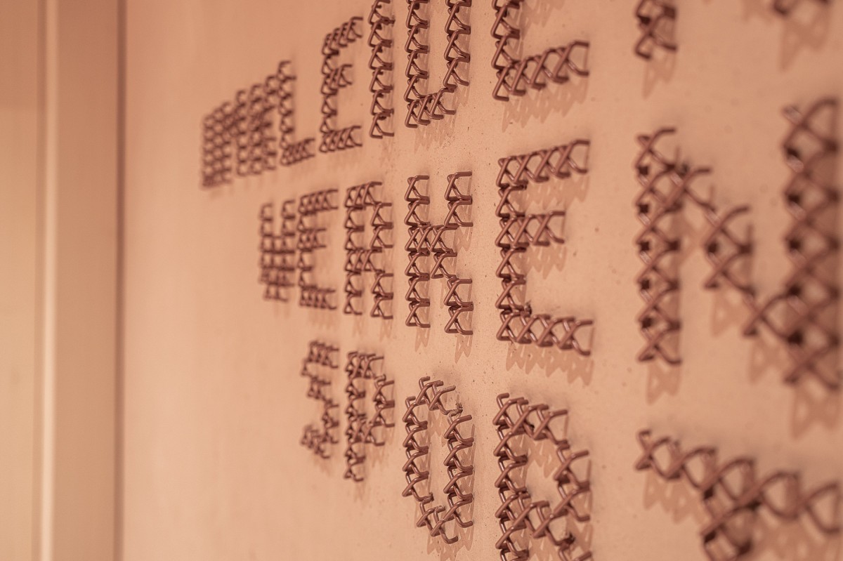 Copper cross-stitch wayfinding letters spelling UMKLEIDEN, WERKEN, SPORT on warm plaster at Education Center Kössen