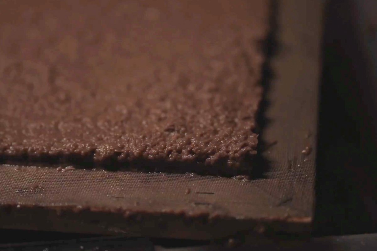 The label material being made: dark sediment paper-pulp spread as a sheet on a deckle frame