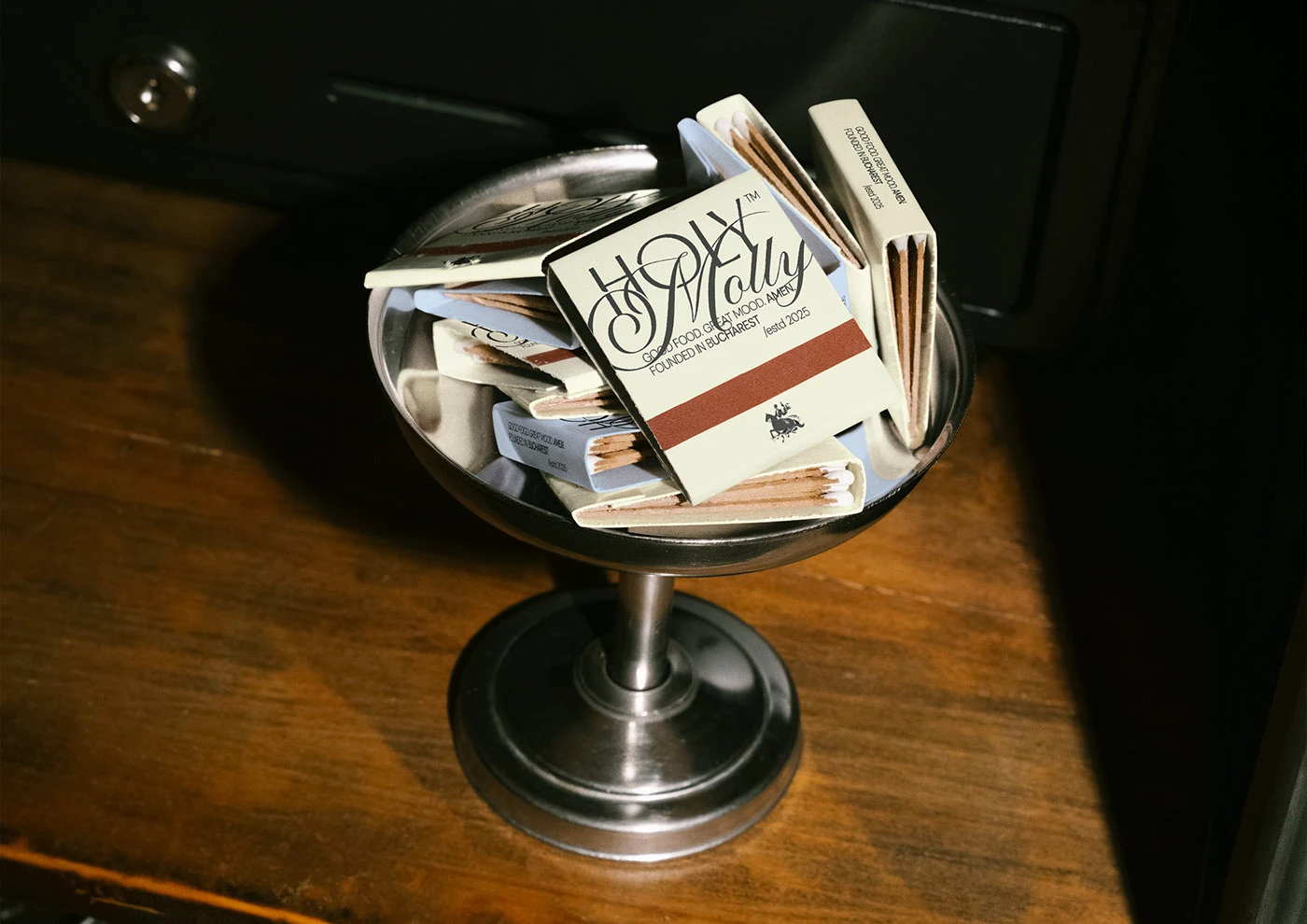 Holy Molly branded matchboxes stacked inside a silver coupe glass, black-and-white on cream — restaurant brand identity