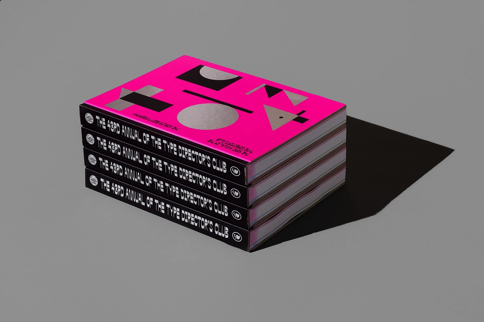 The 43rd Annual of the Type Directors Club hardcover book stack showing fluorescent pink cover, silver foil counterform geometry, and black cloth spines — editorial design by Tereza Bettinardi