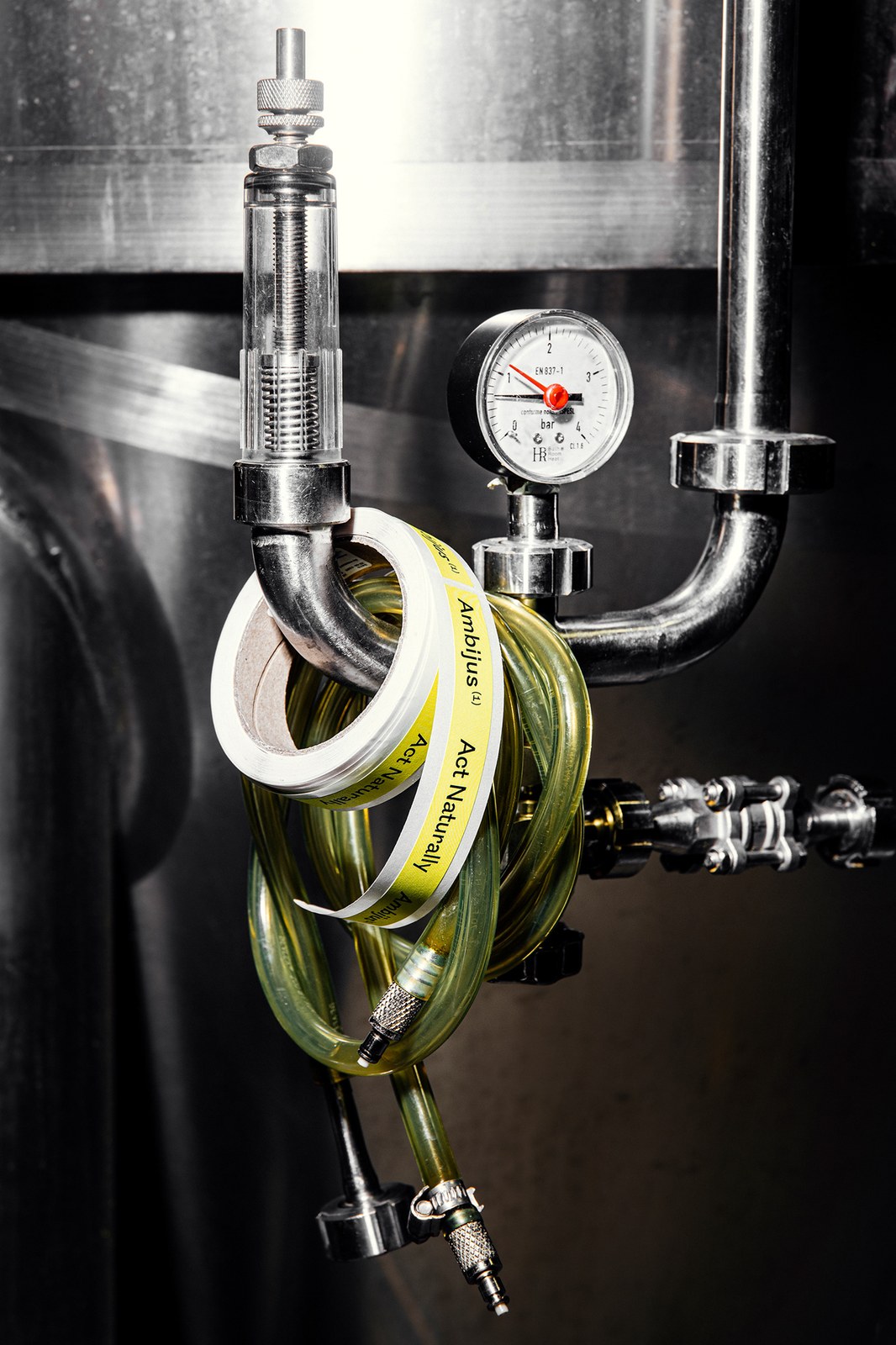 Printed roll of yellow Ambijus Act Naturally neck seal labels hanging on a stainless-steel tank pressure gauge — packaging design by OlssønBarbieri