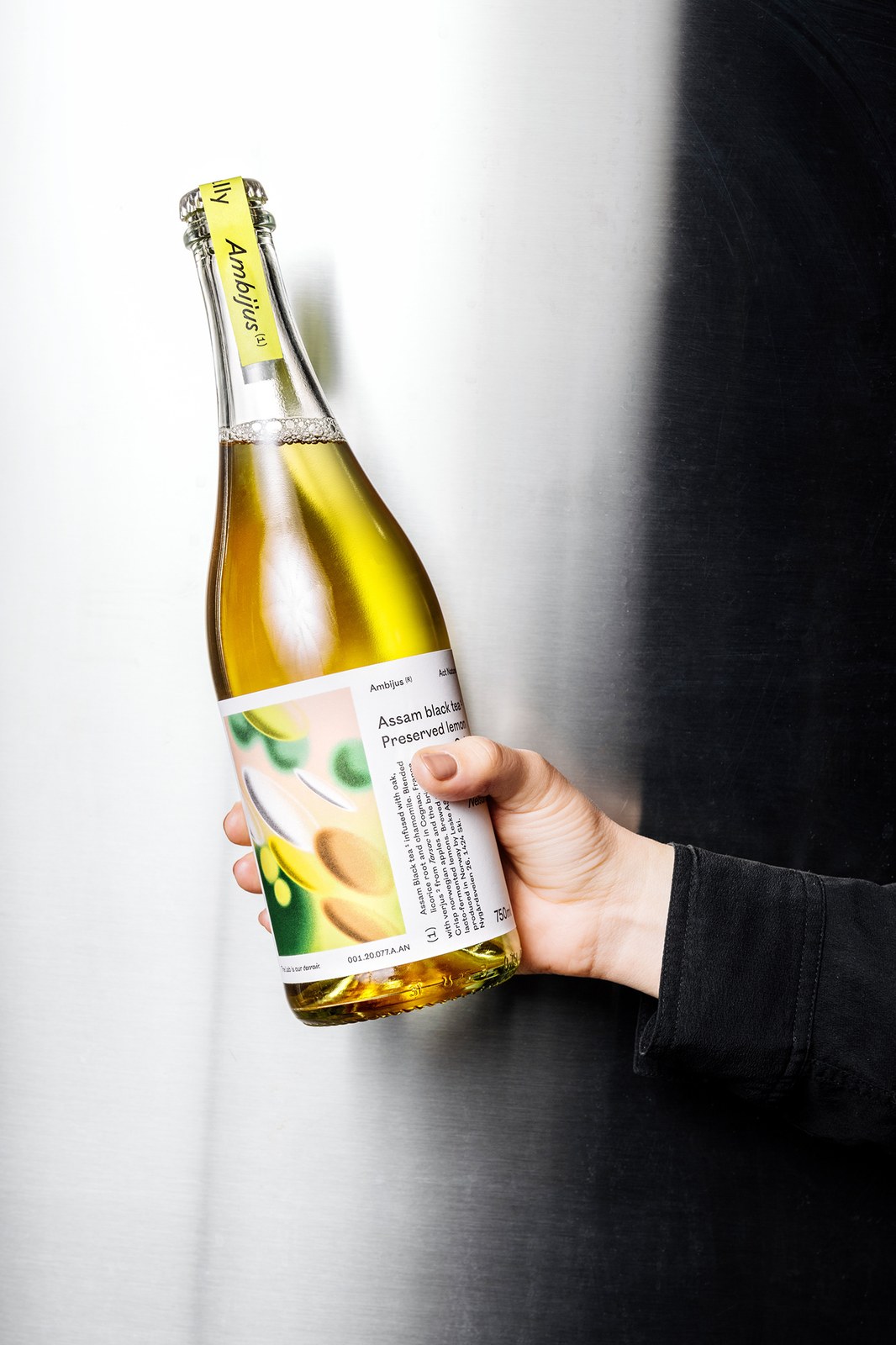 Hand holding Ambijus Act Naturally 750ml glass bottle with uncoated paper label and yellow neck seal — packaging design by OlssønBarbieri
