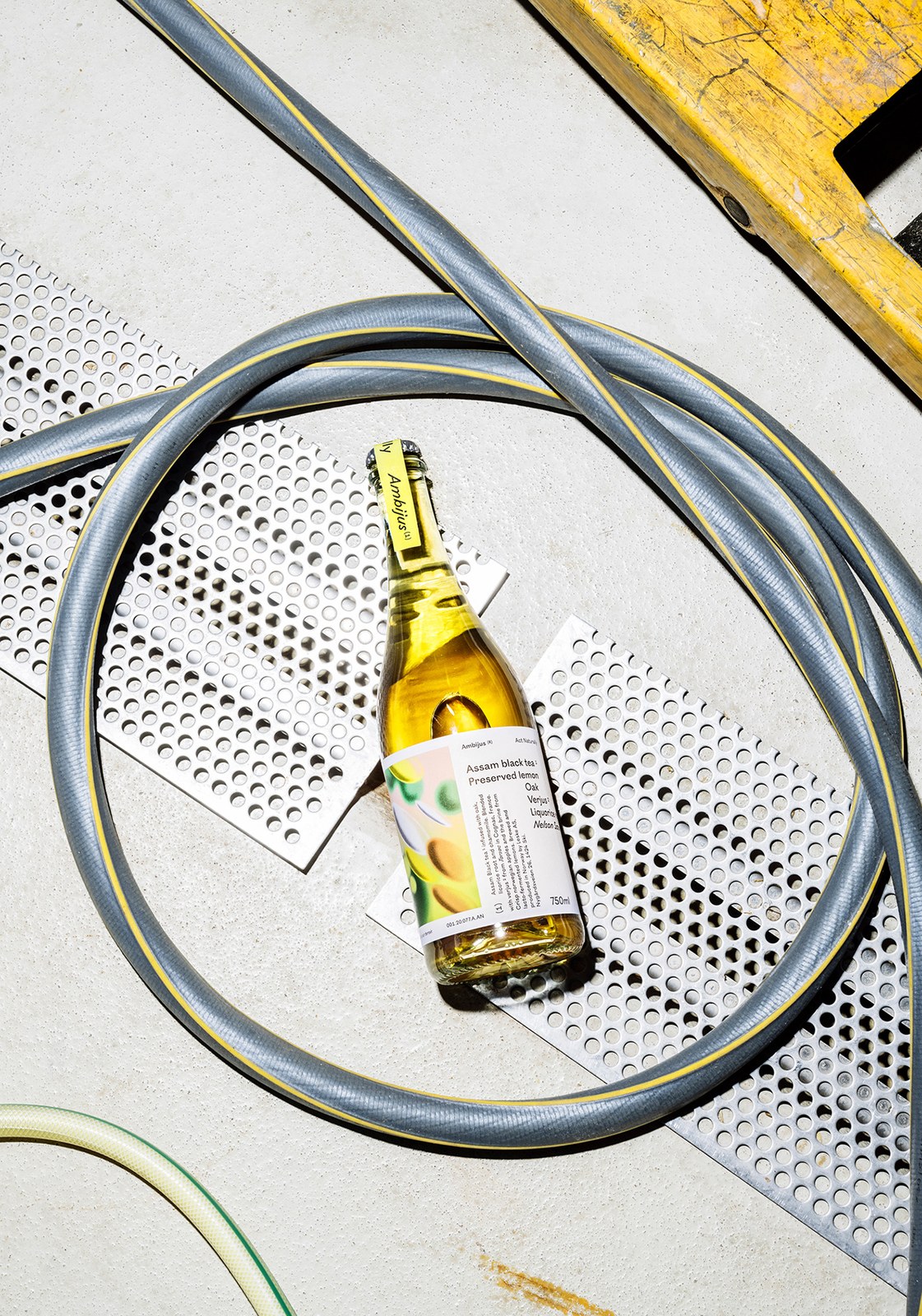 Ambijus Act Naturally bottle resting on perforated steel grates encircled by a coiled brewery hose — packaging design by OlssønBarbieri