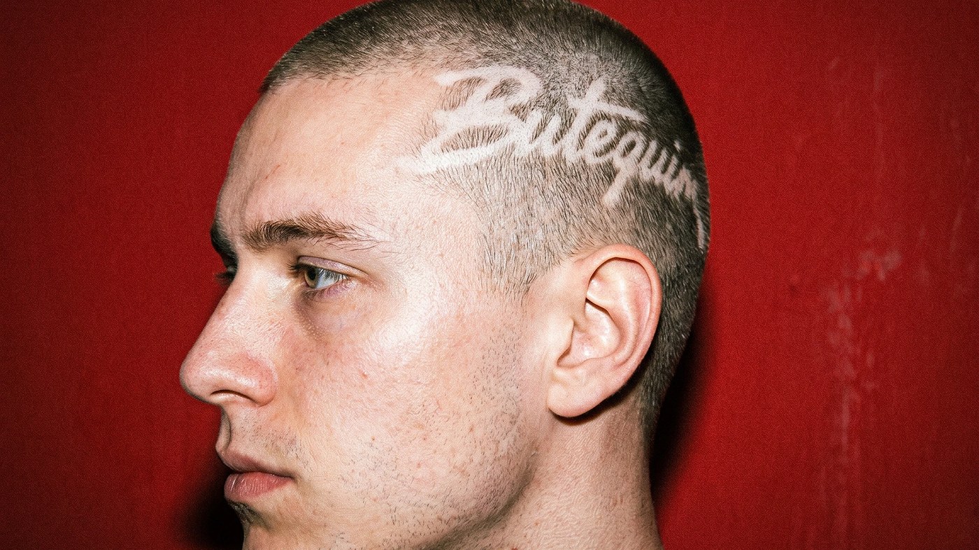 Side profile portrait against red background with Butequim brand identity logotype shaved into buzzcut hair
