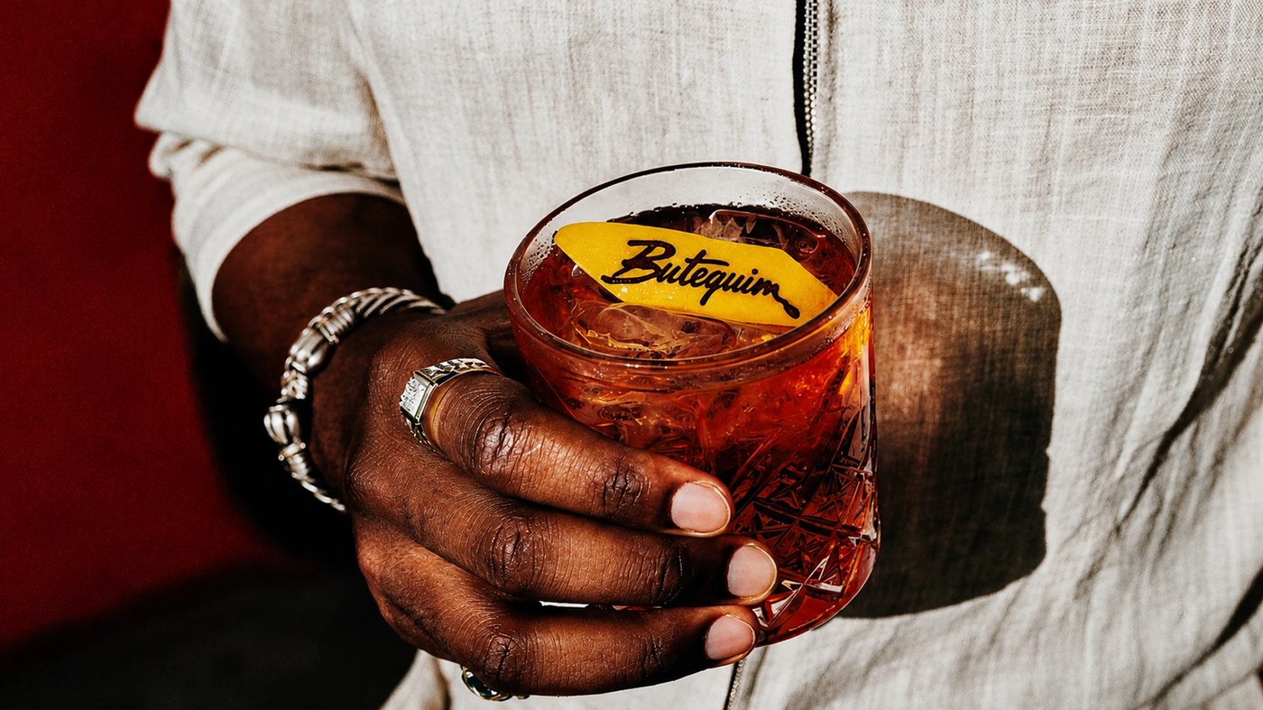 Hand holding cut-crystal rocks glass with branded Butequim citrus peel in brand identity by Estúdio Lippi