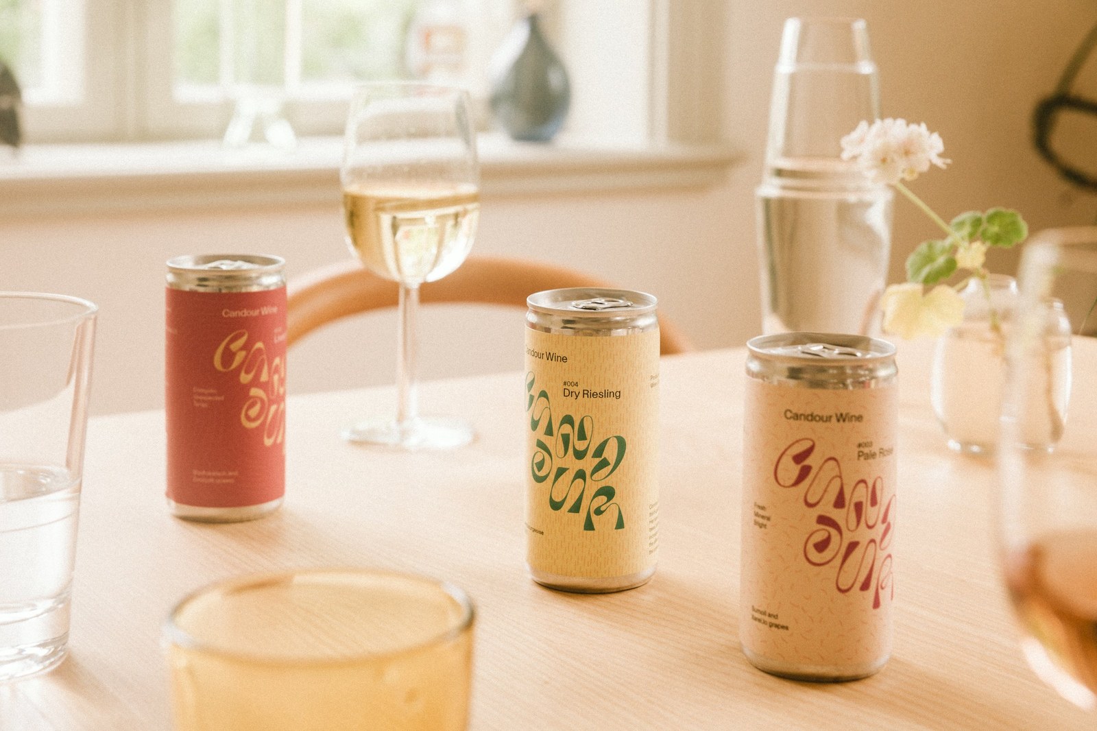 Candour Wine cans arranged on a wooden dining table with white wine glasses by Bedow