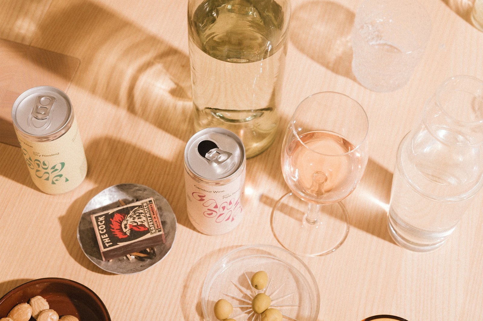 Overhead tabletop view of open Candour Wine #004 Dry Riesling and #003 Pale Rosé cans with stemware and olives