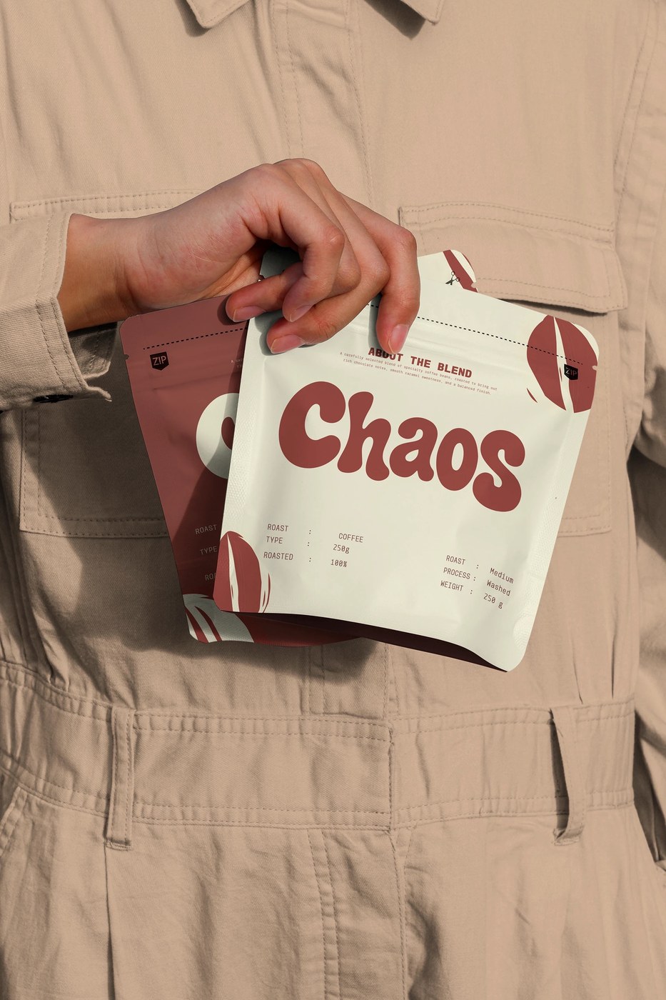 Hands holding terracotta Chaos coffee pouches against a khaki jumpsuit displaying roast telemetry and bold typography