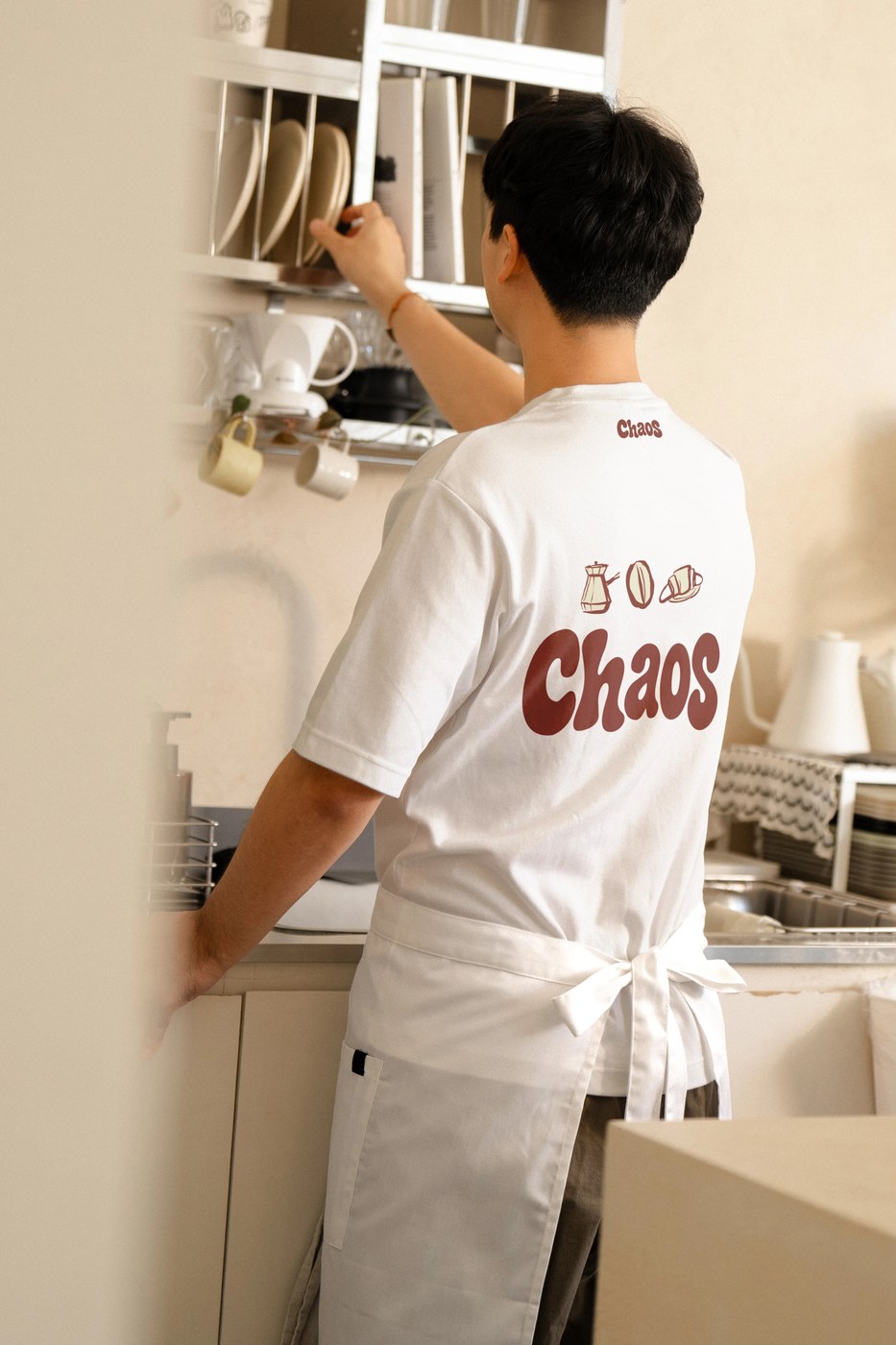 Barista reaching toward cafe shelving in black apron and custom screenprinted staff t-shirt featuring the large Chaos wordmark across the back