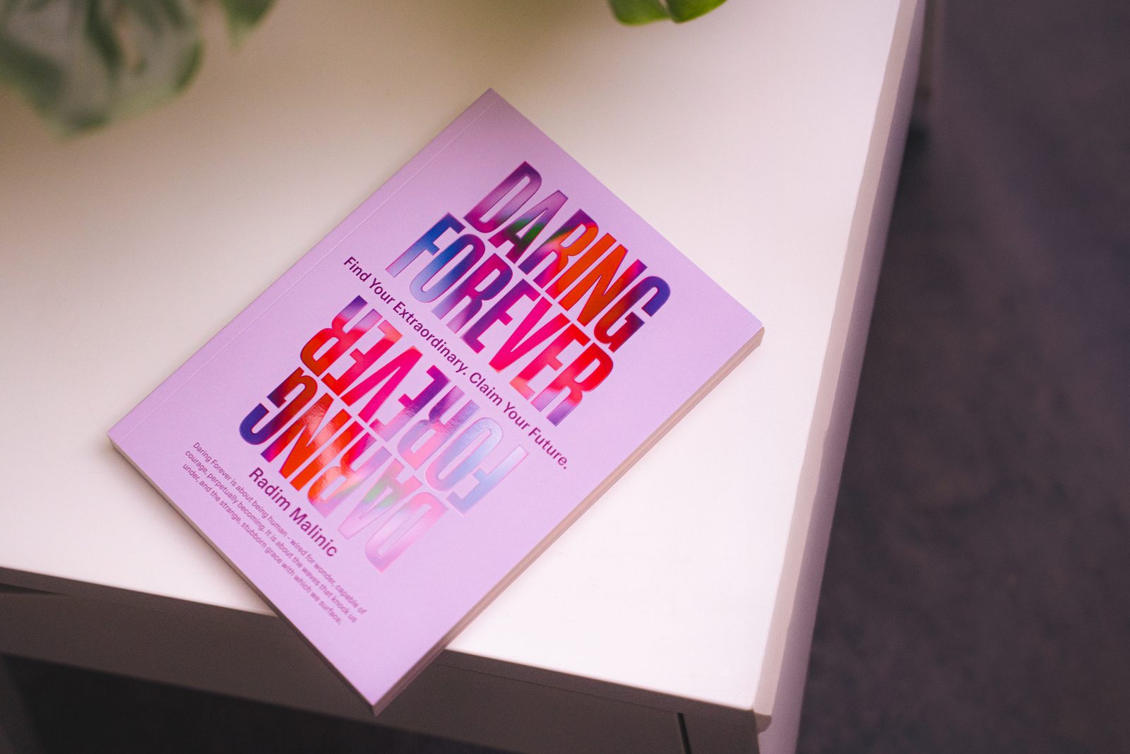 Paperback cover resting on a white table beside a plant, Daring Forever book by Radim Malinic