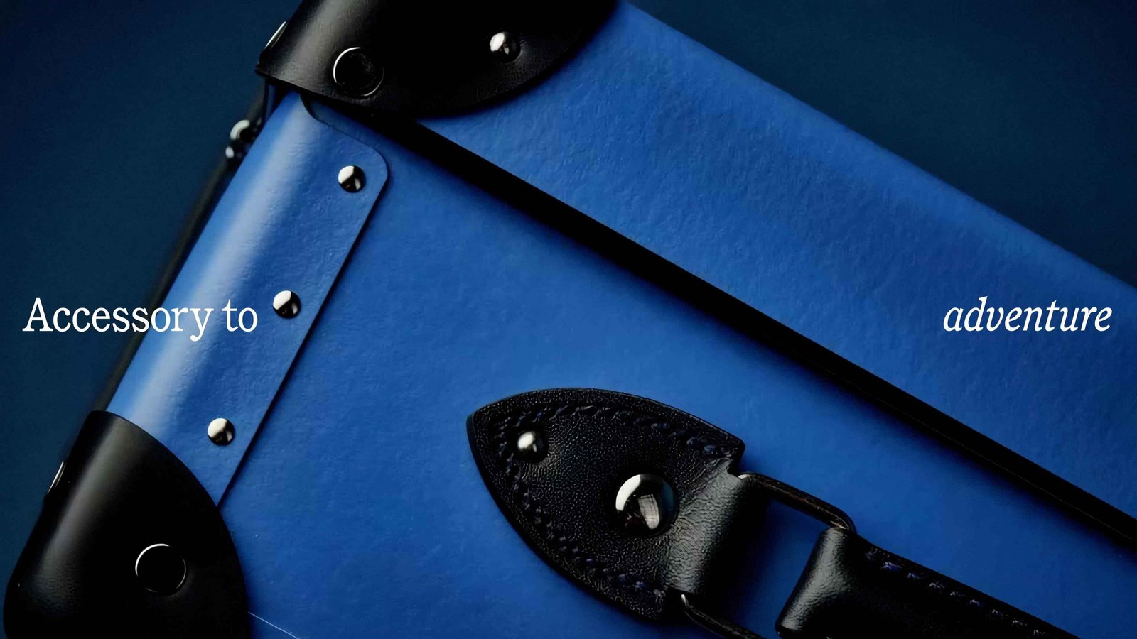 Close-up of a cobalt-blue vulcanised fibreboard case with black leather corners, silver rivets, and Accessory to adventure serif headline — brand identity by Ragged Edge