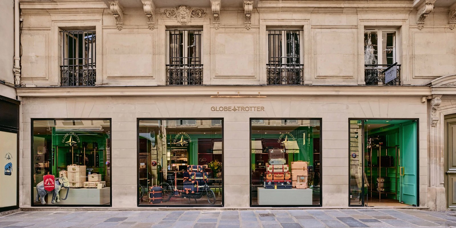 Paris flagship storefront exterior featuring brass signage, emerald-green interior walls, stacked trunks, and the heritage elephant display — brand identity by Ragged Edge