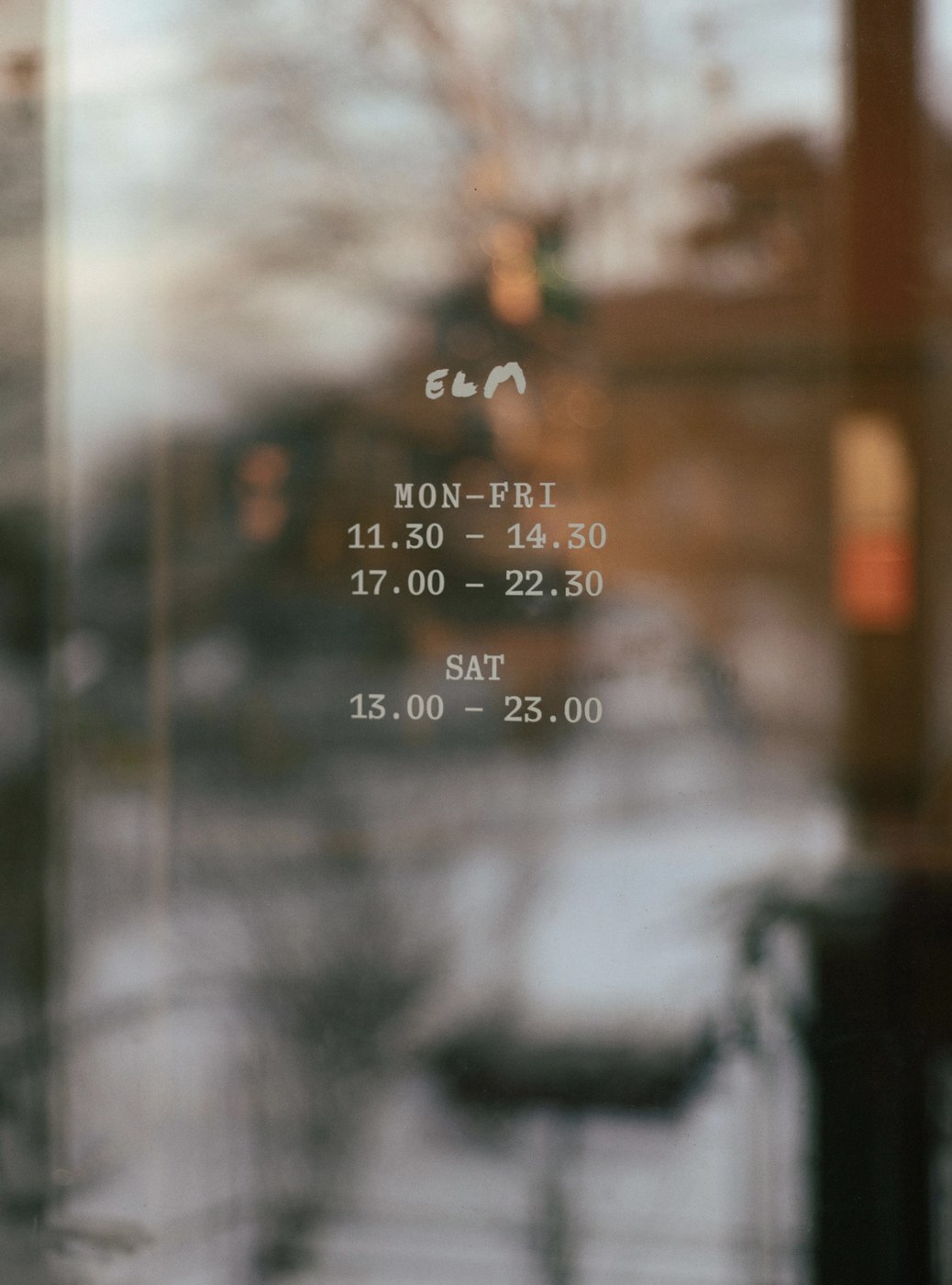 Glass entrance door featuring white vinyl brushstroke Elm logo and opening hours