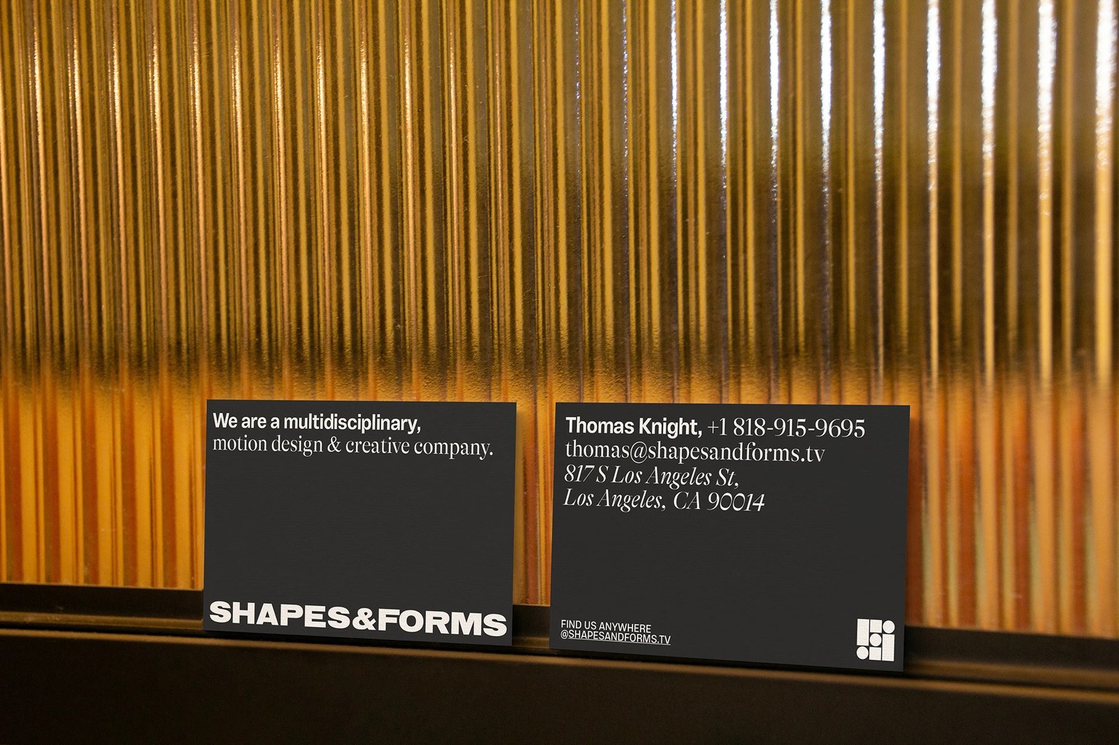 Shapes & Forms matte-black business cards leaning against warm amber fluted glass