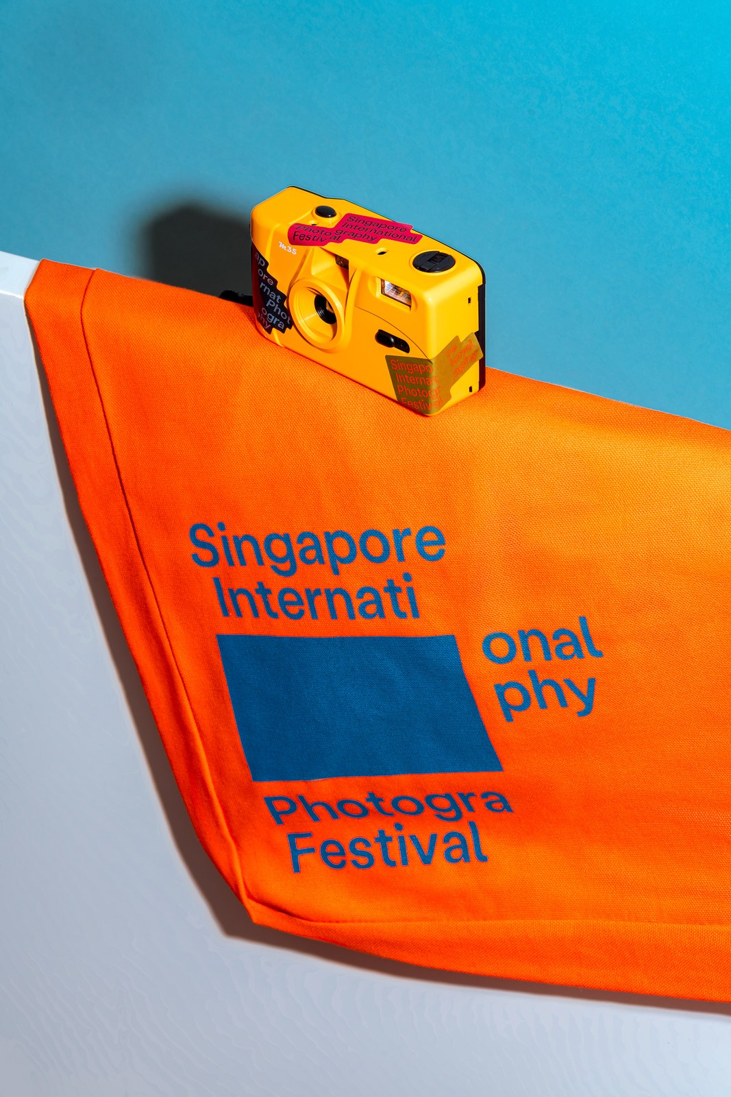 Orange canvas SIPF tote bag screenprinted in cobalt blue beneath a yellow 35mm camera covered in typographic stickers