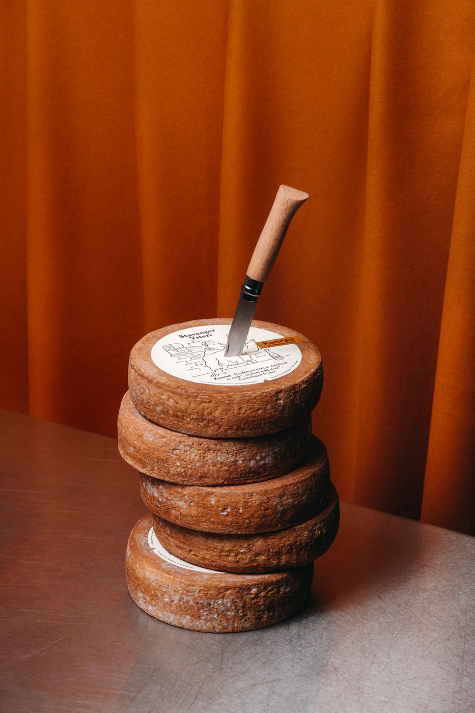 Stack of Konrad cheese wheels with Opinel knife in top wheel, featuring circular label and brown name tag in packaging design by OlssønBarbieri
