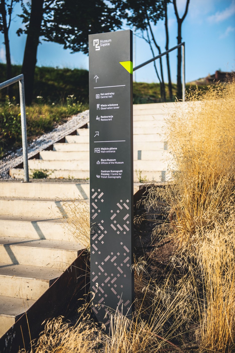 Vertical wayfinding totem with bilingual typography, custom pictograms, and lime green directional accent in spatial design