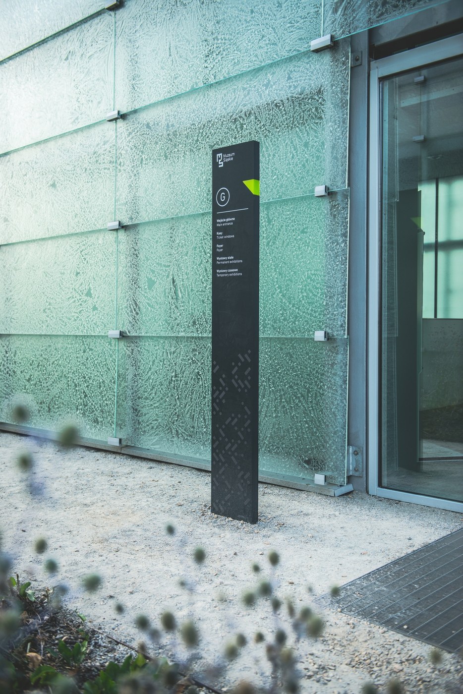 Level G gallery entrance totem set against textured translucent glass facade in architectural spatial design